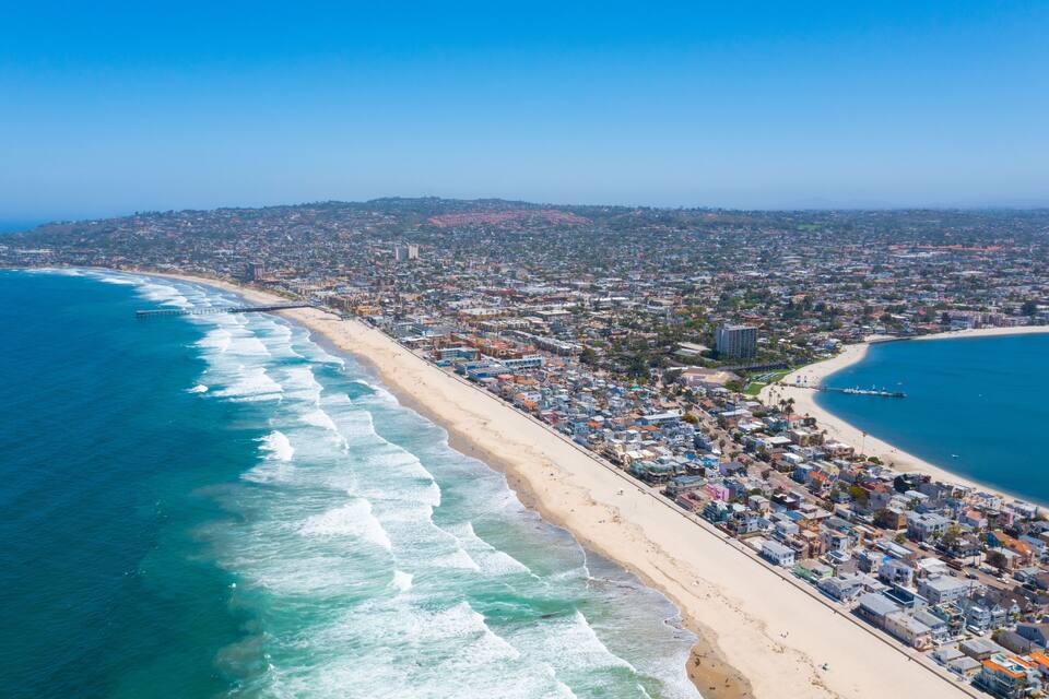A coastal cityscape with a long sandy beach, a pier, and dense urban buildings.