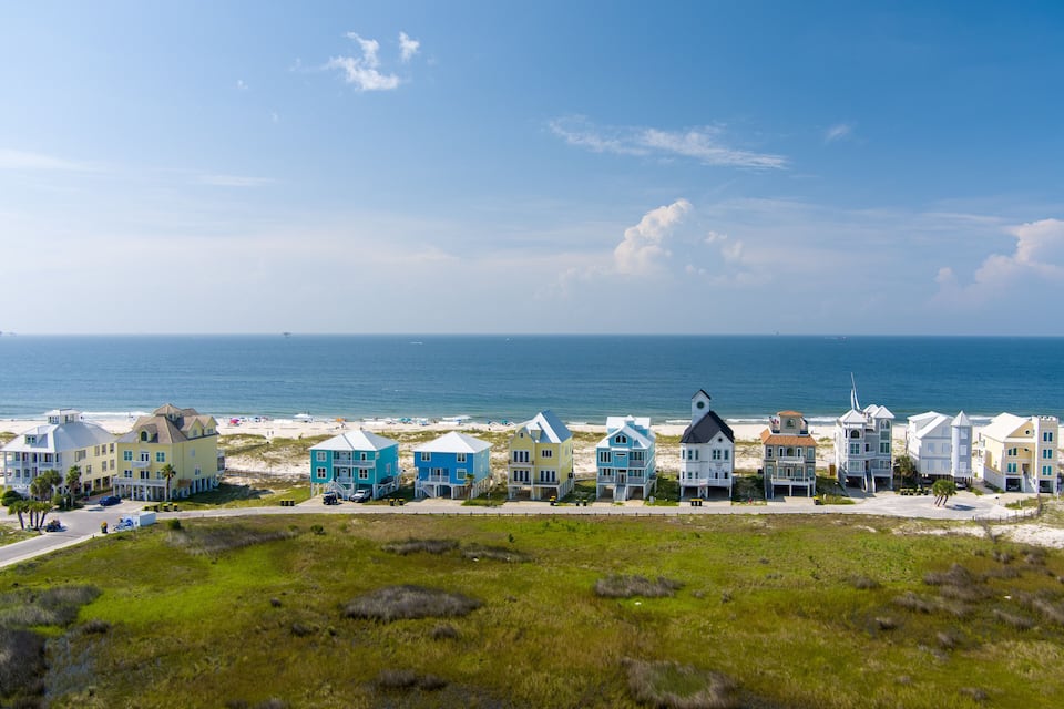 Een kustgebied met woonhuizen in verschillende kleuren langs het strand, een helderblauwe lucht en de oceaan op de achtergrond.
