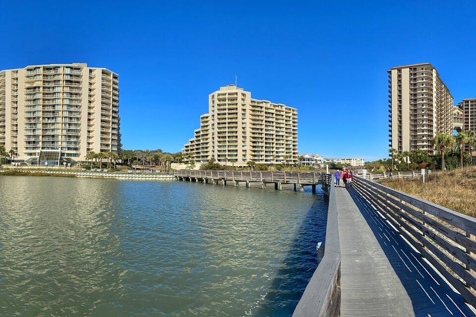 A waterfront promenade with high-rise buildings, a wooden walkway, and a clear blue sky.