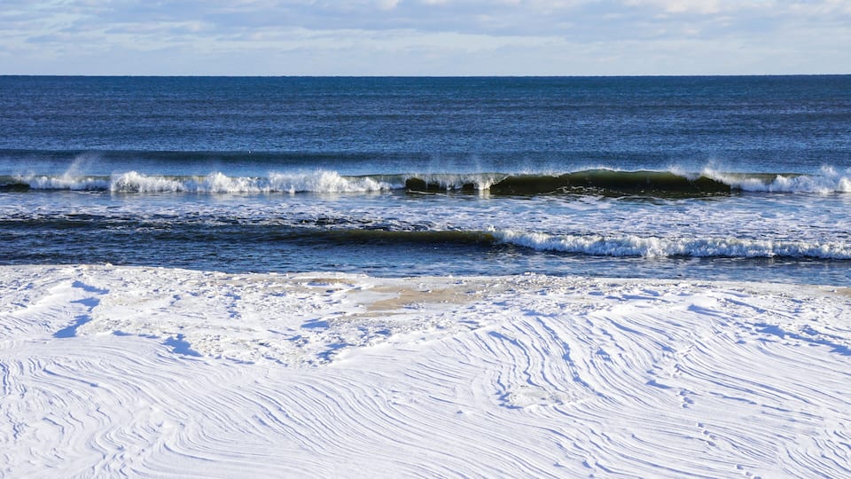 A snowy beach with waves crashing onto the shore and a clear blue sky.