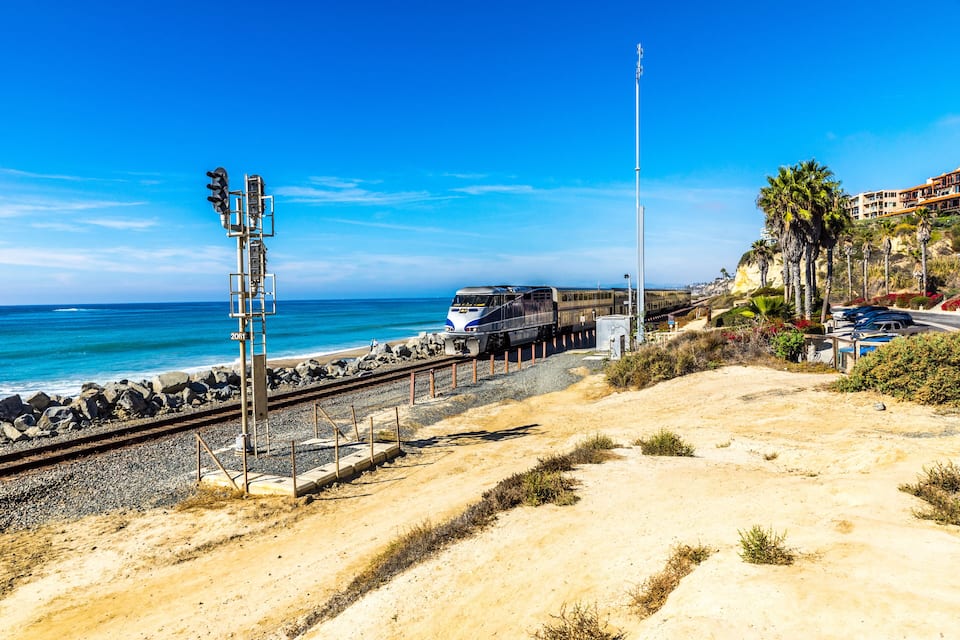 A coastal train track with a train, beach, and palm trees.