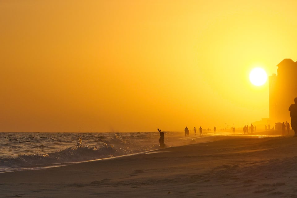 A beach at sunset with people walking along the shore and a building in the background.