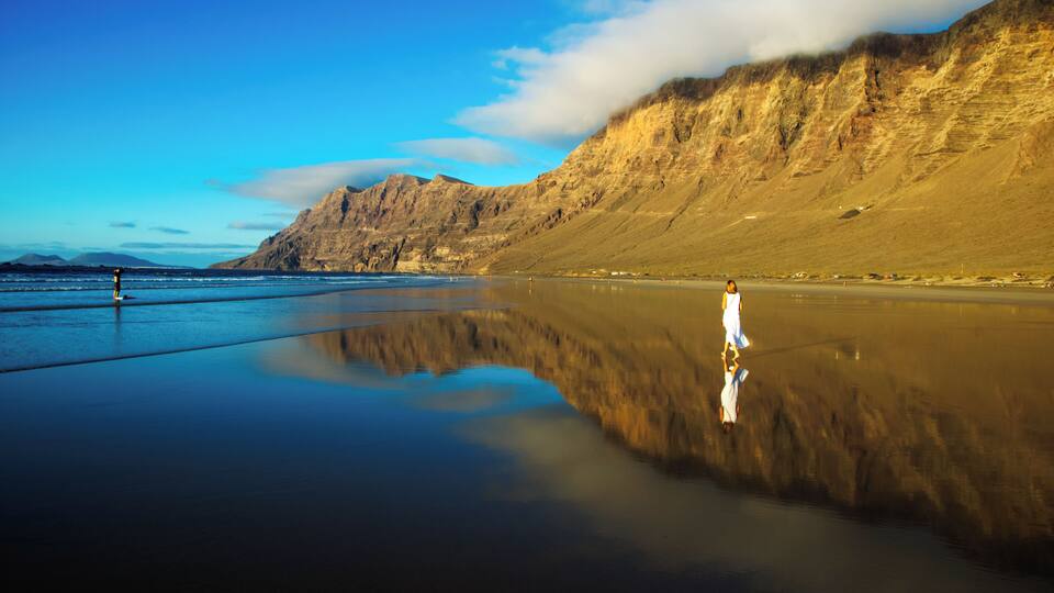A beach with a person standing on the sand, mountains in the background, and a clear sky.