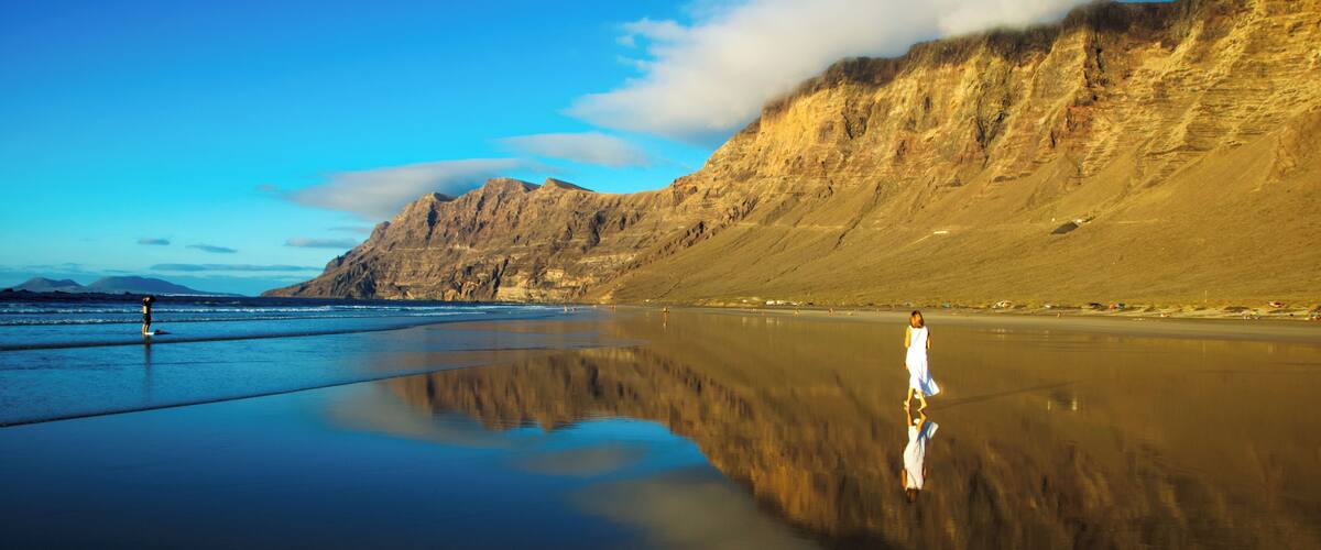 A beach with a person standing on the sand, mountains in the background, and a clear sky.