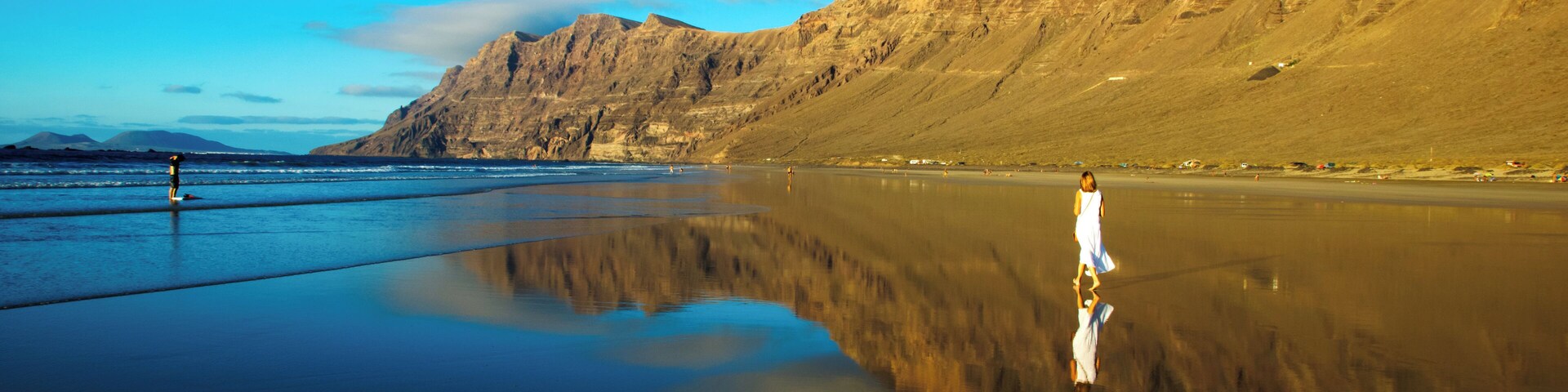 A beach with a person standing on the sand, mountains in the background, and a clear sky.