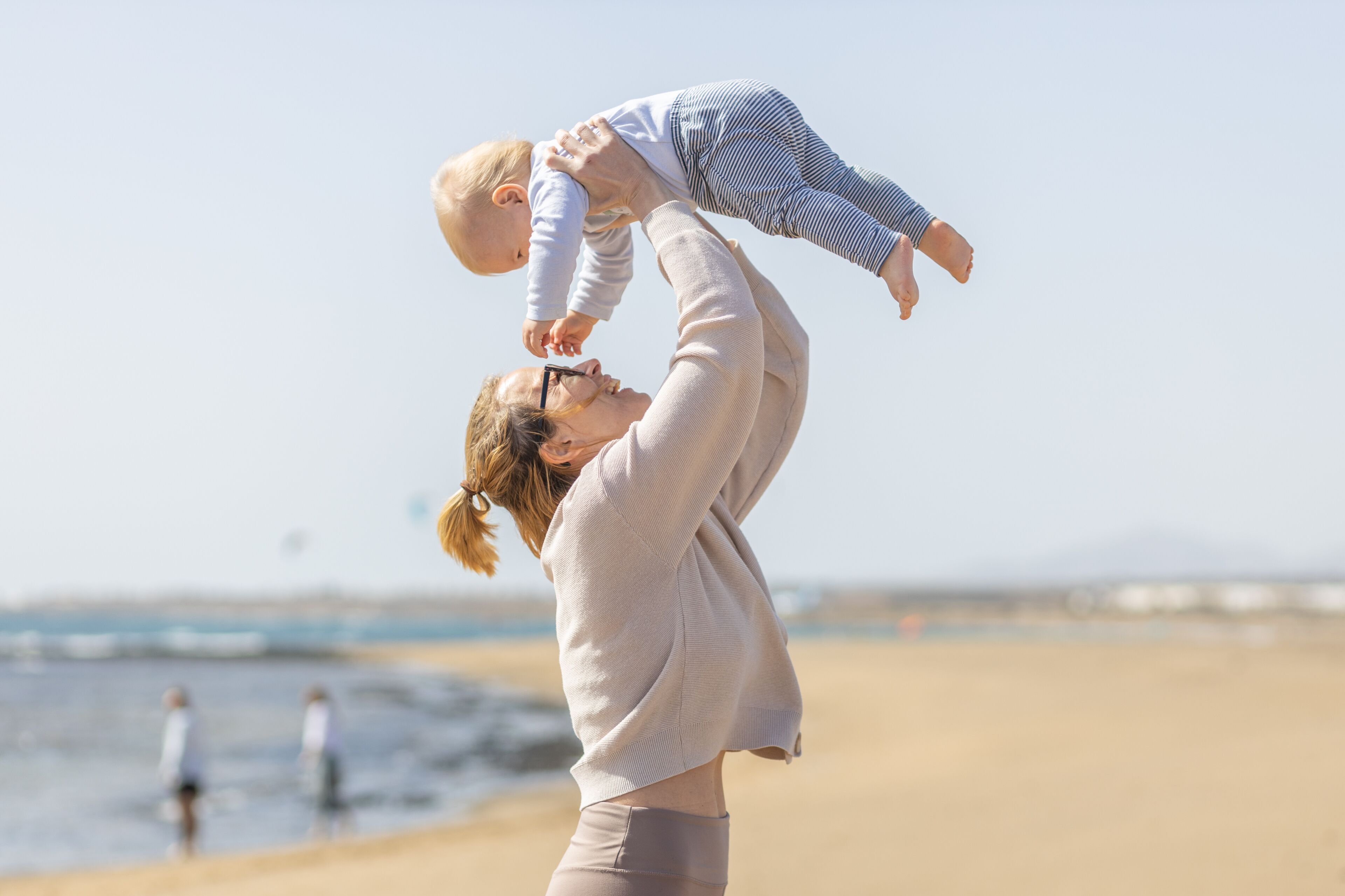 A beach scene with a person holding a child in the air, a sandy shore, and the ocean in the background.