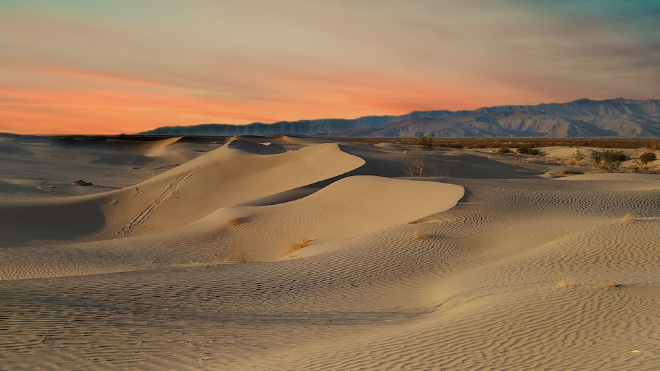Sand dunes with a sunset sky and distant mountains.
