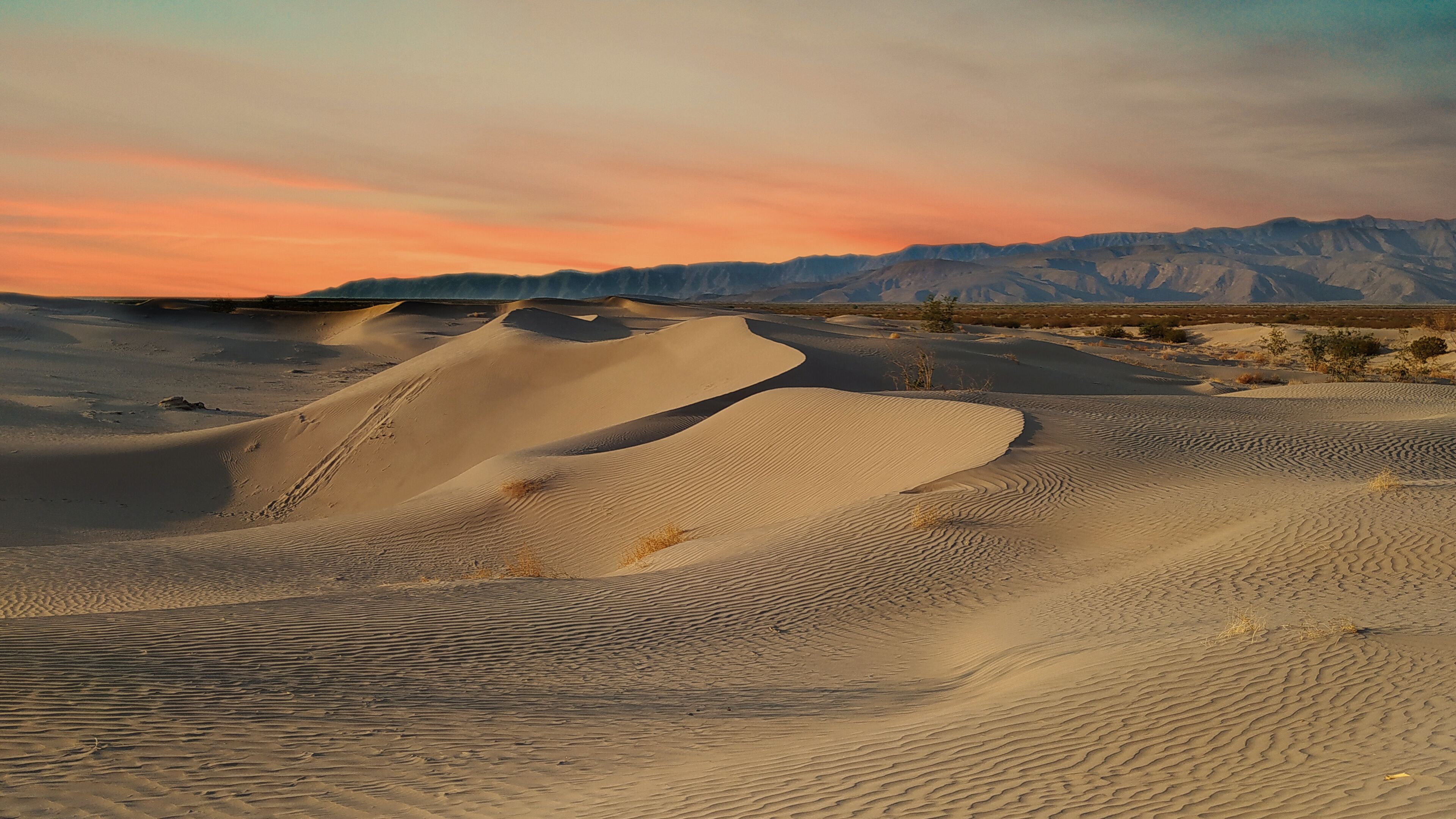 Sand dunes with a sunset sky and distant mountains.