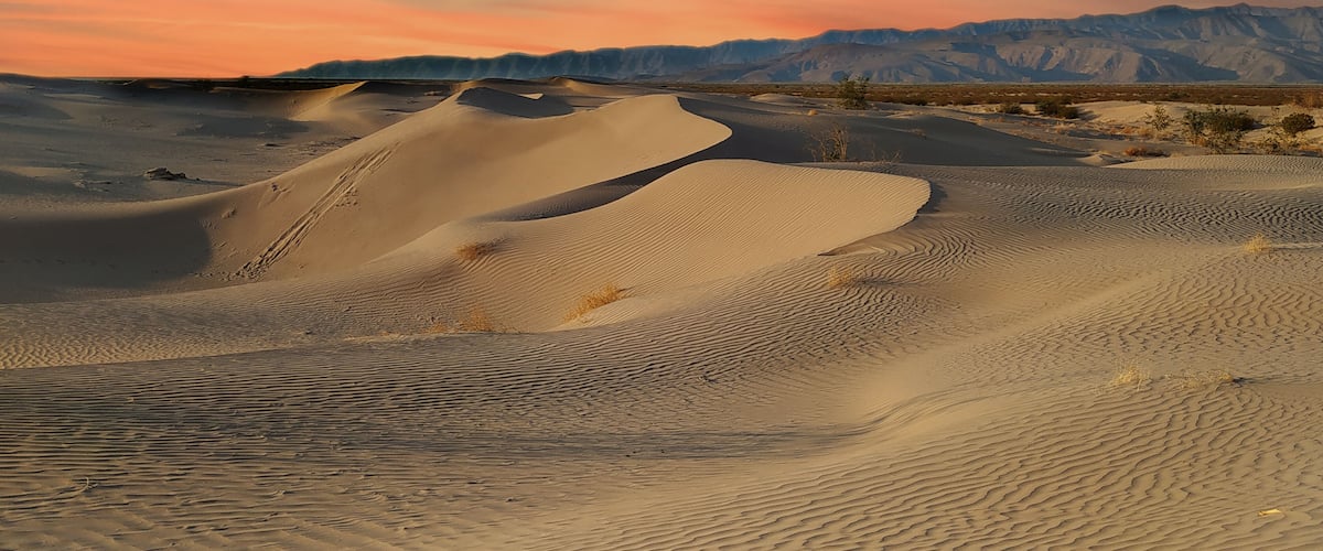 Sand dunes with a sunset sky and distant mountains.