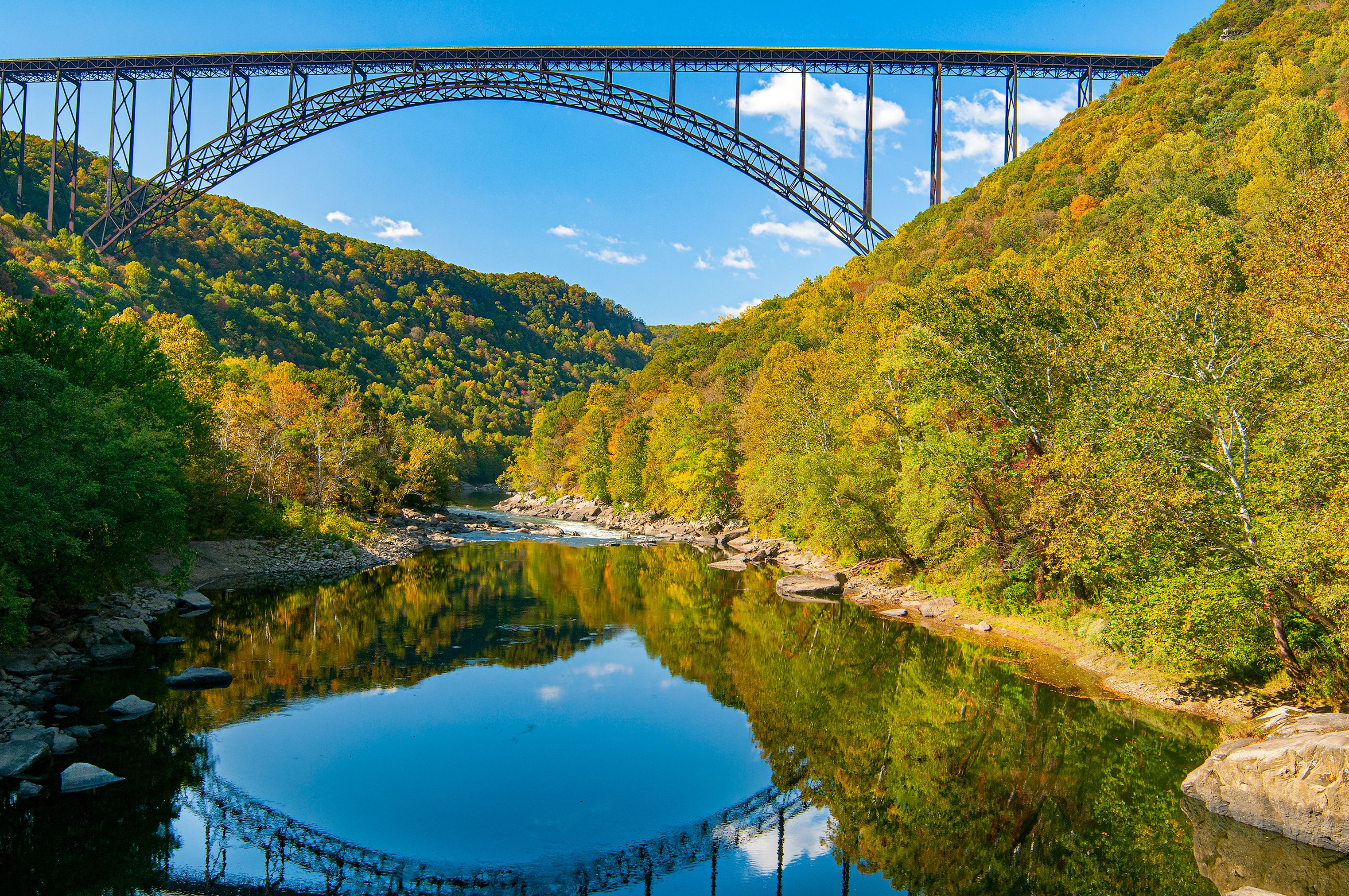 A steel bridge arching over a calm river, surrounded by lush green hills and reflecting the sky.