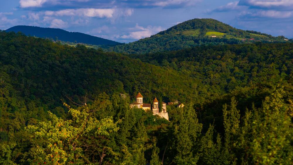 A lush green landscape with a historic building nestled among trees, a hill with a small structure on top, and a distant mountain range under a partly cloudy sky.
