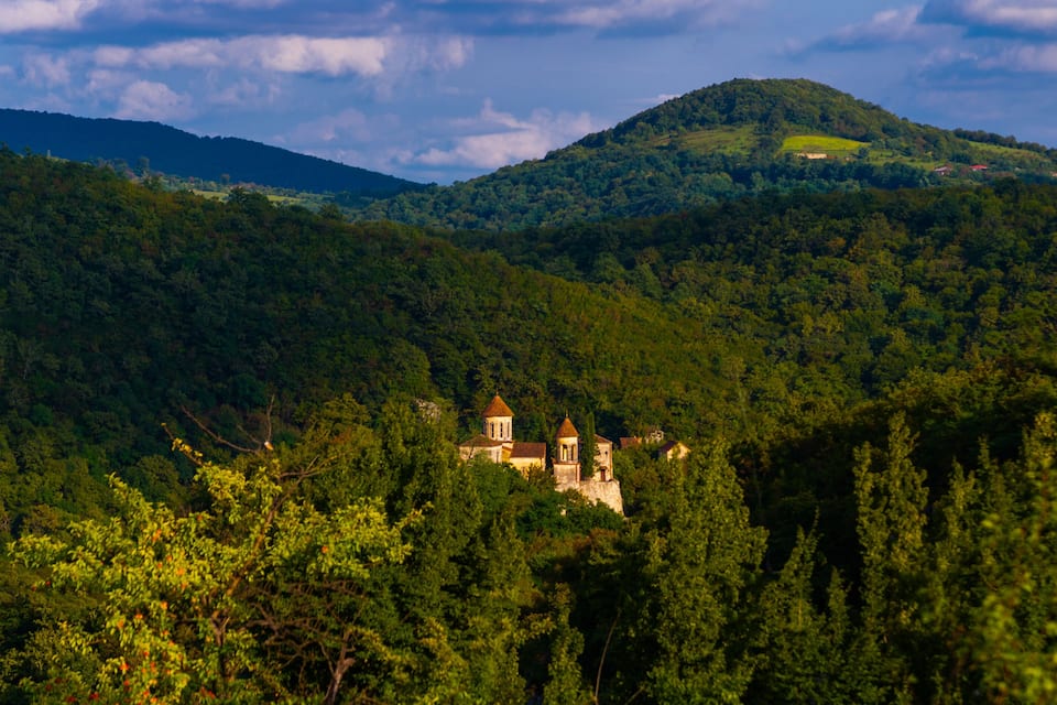 A lush green landscape with a historic building nestled among trees, a hill with a small structure on top, and a distant mountain range under a partly cloudy sky.