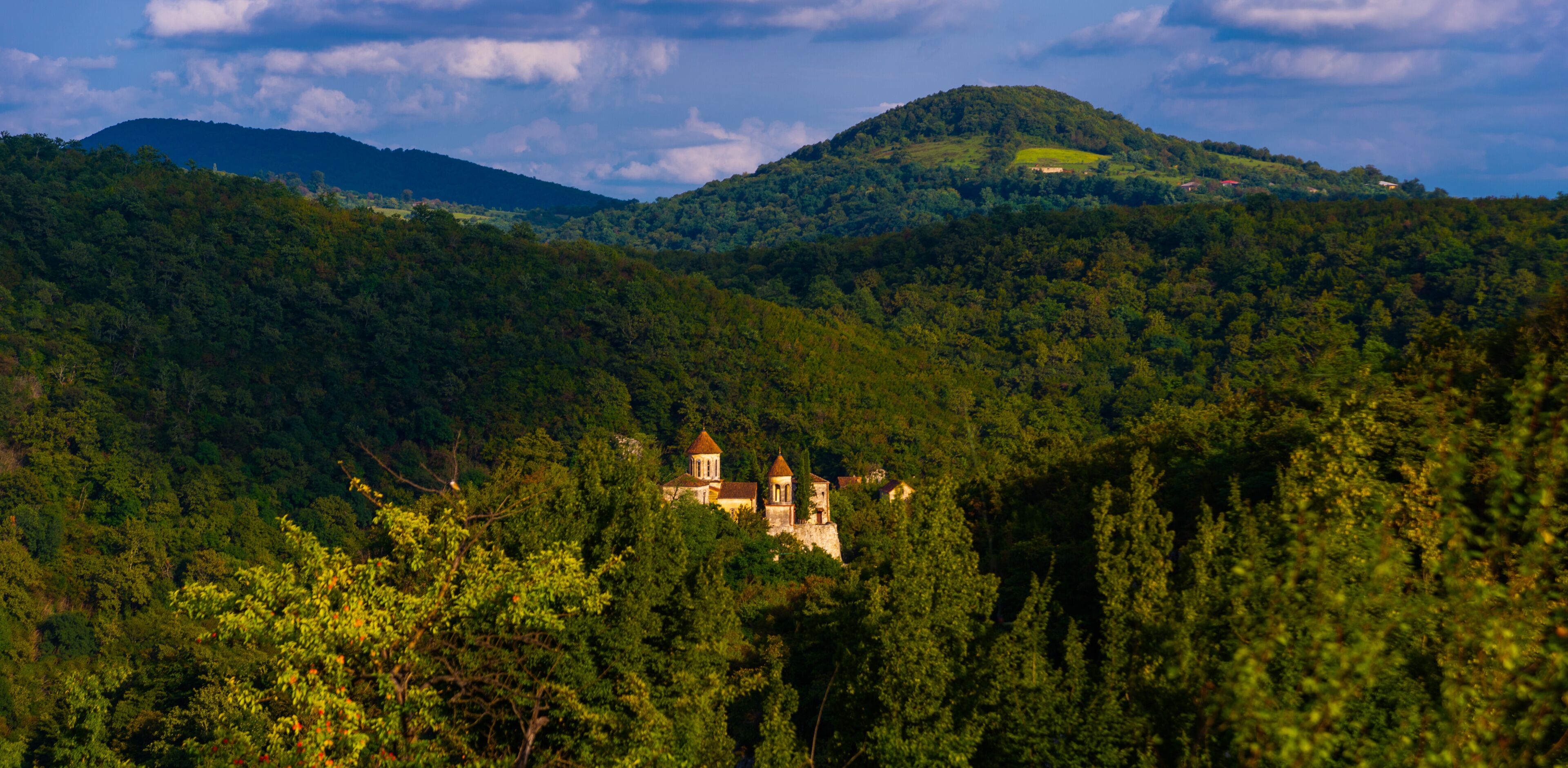 A lush green landscape with a historic building nestled among trees, a hill with a small structure on top, and a distant mountain range under a partly cloudy sky.