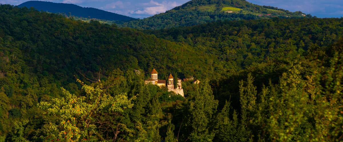 A lush green landscape with a historic building nestled among trees, a hill with a small structure on top, and a distant mountain range under a partly cloudy sky.