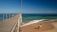 A wooden pier extending into the ocean, with people walking along it and a couple walking on the beach.