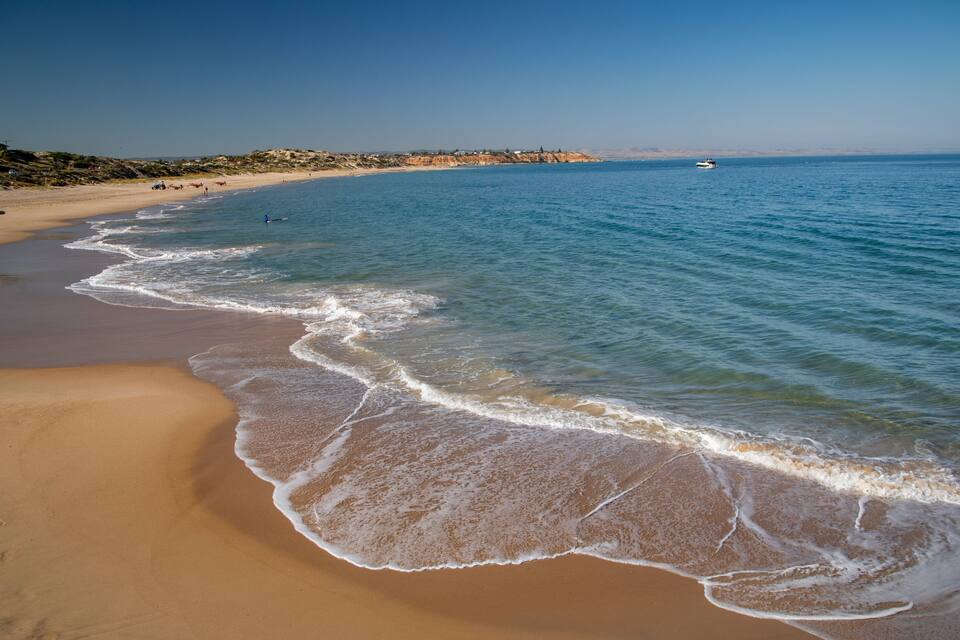 Uma praia de areia com ondas suaves, céu azul límpido e um barco ao longe na água.