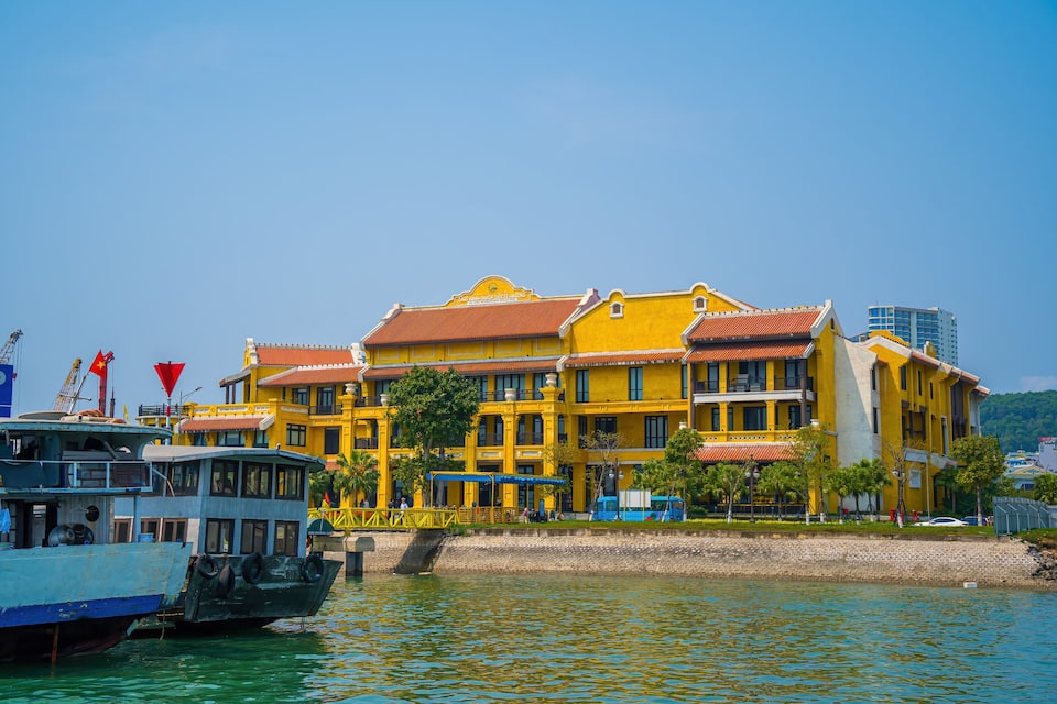 A waterfront building with a yellow facade and red roof, boats docked nearby, and a clear blue sky.