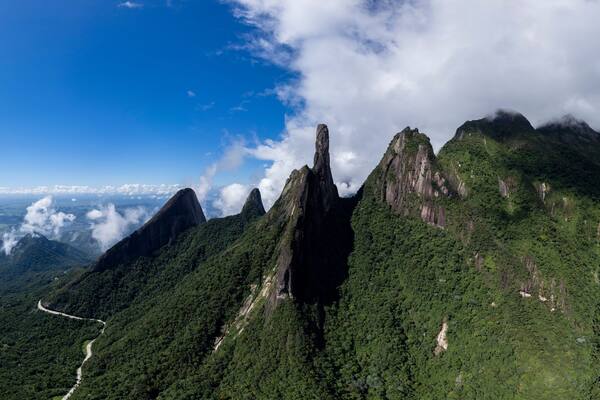 A mountainous landscape with a winding road, dense green forests, and prominent rock formations under a clear blue sky.