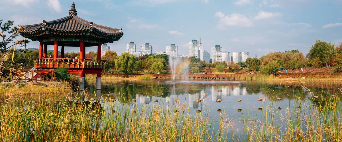 A traditional pavilion by a calm lake with a city skyline in the background.