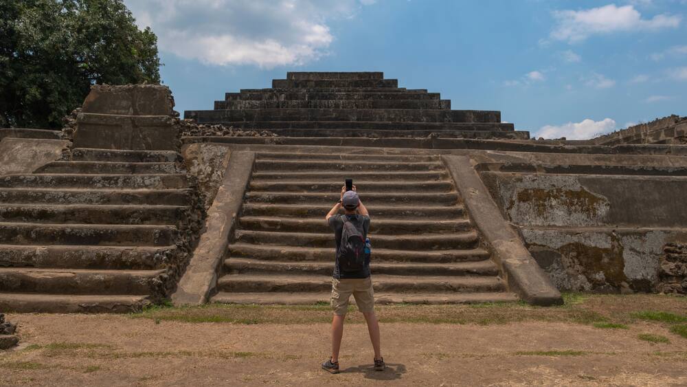 Young man tourist taking photos with the phone at the main pyramid with temple at Tazumal Park archaeological site in El Salvador.