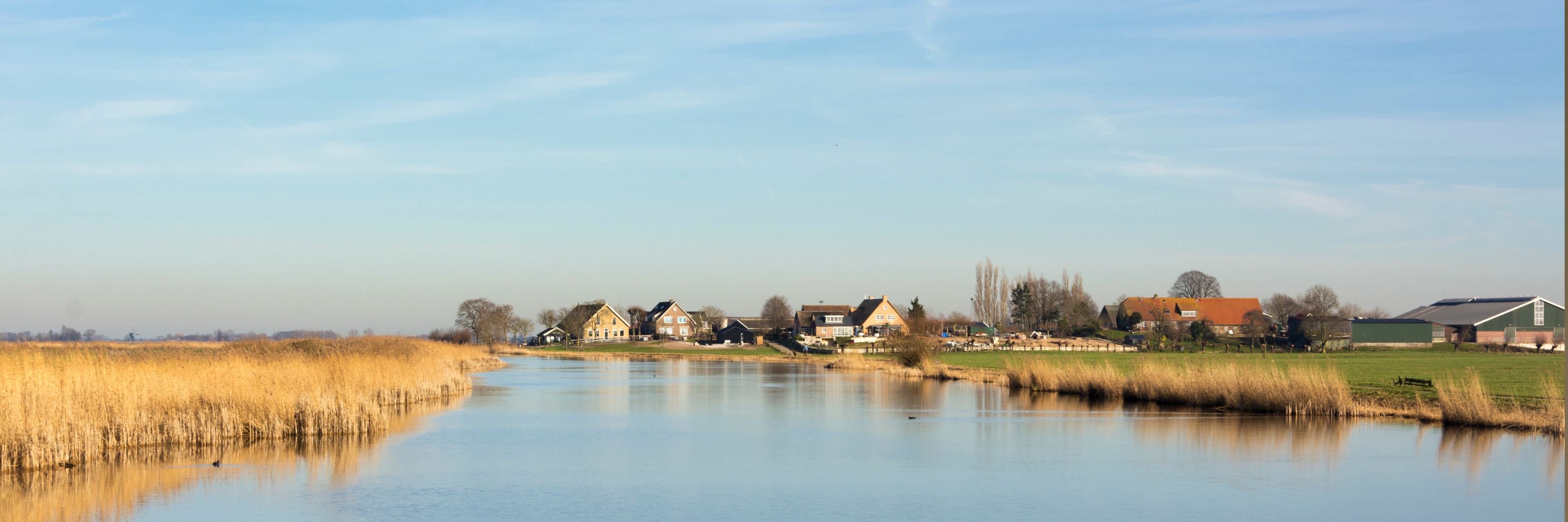 A river with houses along its banks, a clear sky, and a calm water surface reflecting the surroundings.