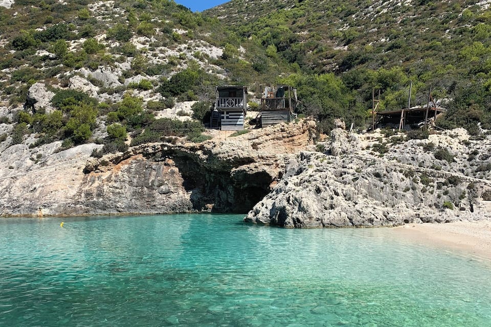 A clear turquoise sea with a rocky shore, a red boat, and a small wooden structure on a hill.