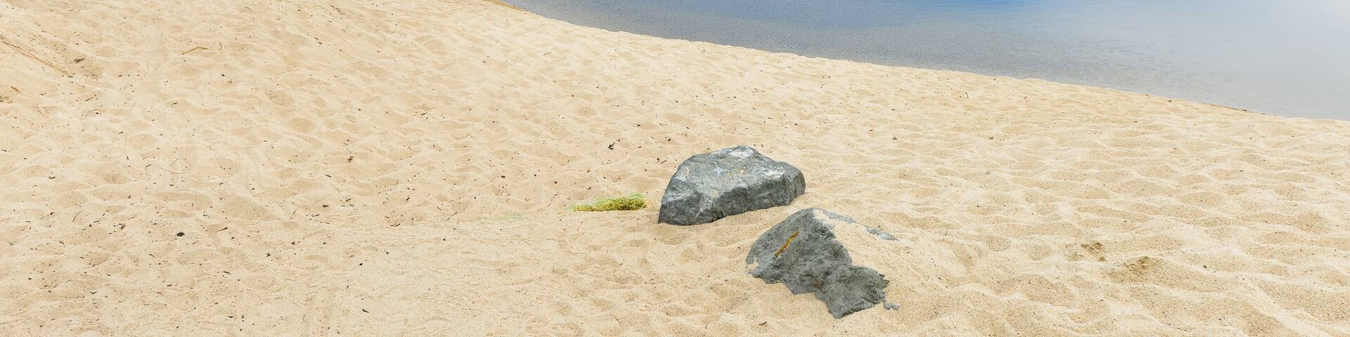 A riverbank with sand and rocks, a calm river, and a mountainous landscape in the background.