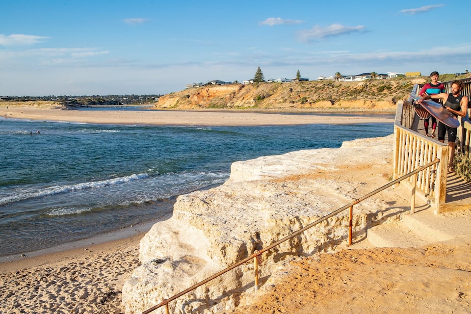 A coastal beach with a rocky shoreline, a wooden boardwalk, and houses in the background.