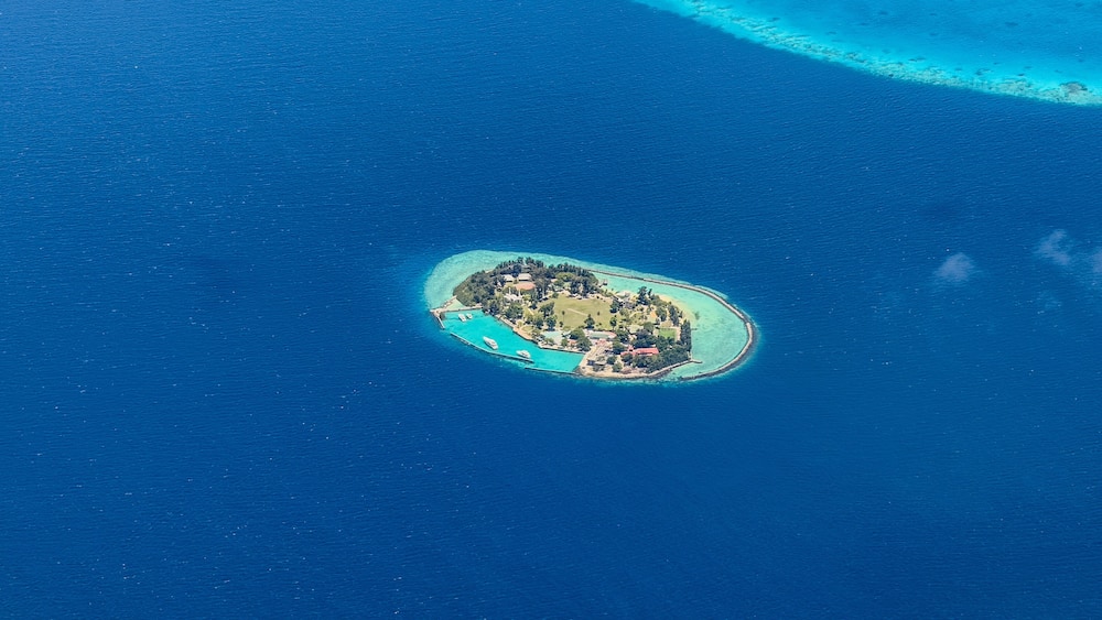 aerial landscape view of Aarah island as part of Kaafu Atoll with buildings, trees, other infrastructure and harbor, surrounded by deep blue water
