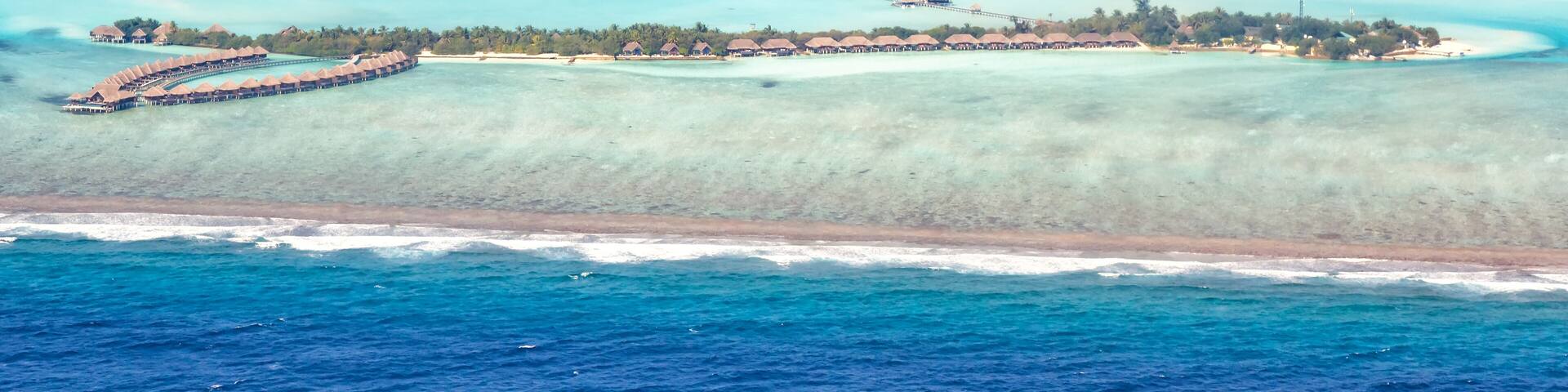An aerial view of a tropical island with clear turquoise waters, a long stretch of beach, and a resort area with overwater bungalows.