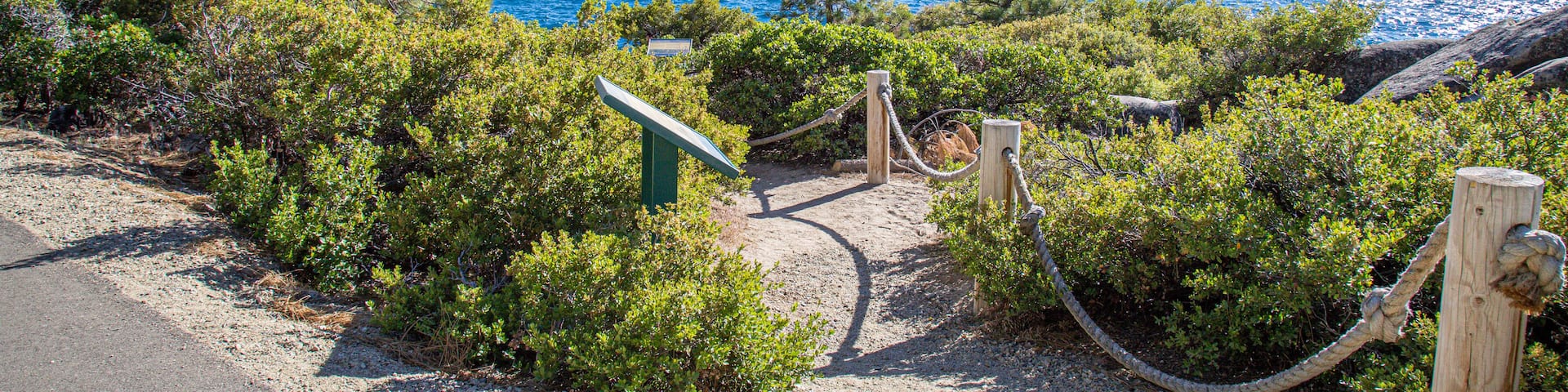 A coastal trail with a wooden sign, dense greenery, and a clear view of the ocean and distant mountains.