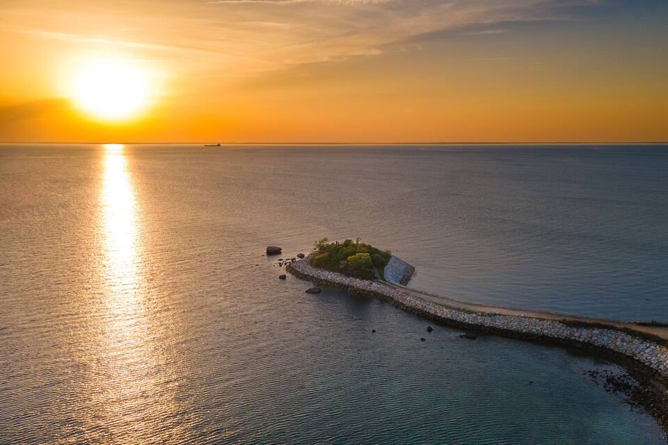 Un sol ponent sobre un mar tranquil amb una petita illa connectada per un drom de pedra.