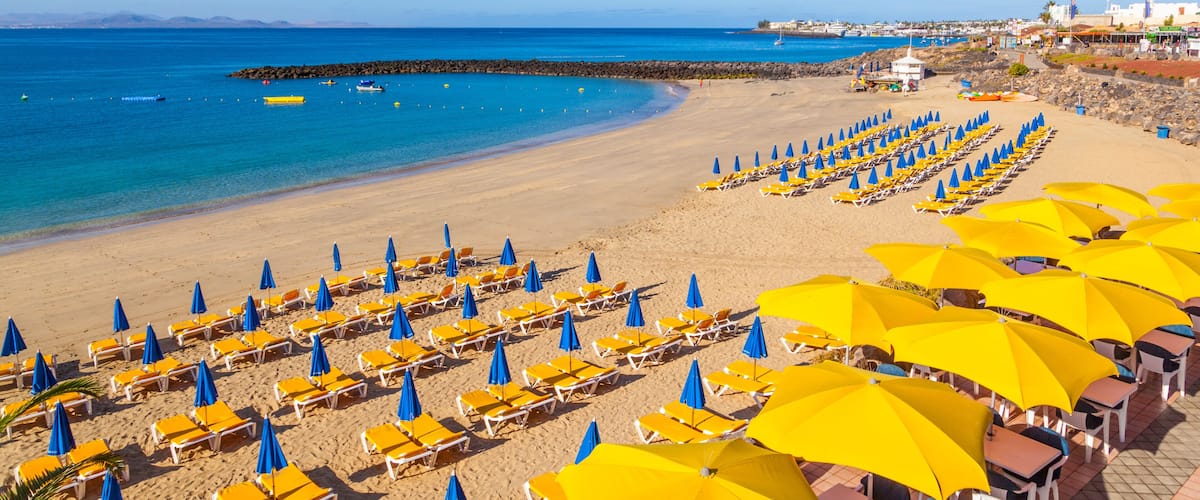 A beach with rows of yellow and blue umbrellas, a rocky breakwater, and a clear blue sea.
