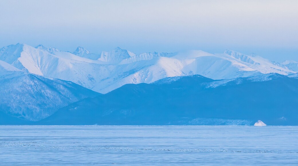 View of the mountains on the coast of ice-covered Lake Baikal. Winter panoramic landscape. Snow-capped mountain peaks in the distance. Evening panorama. Republic of Buryatia, Siberia, Russia.