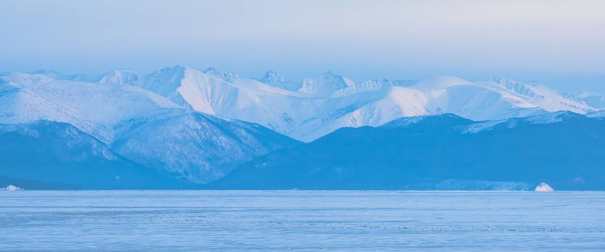 View of the mountains on the coast of ice-covered Lake Baikal. Winter panoramic landscape. Snow-capped mountain peaks in the distance. Evening panorama. Republic of Buryatia, Siberia, Russia.