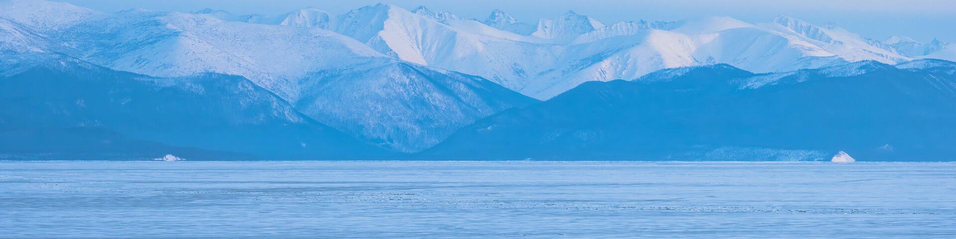 View of the mountains on the coast of ice-covered Lake Baikal. Winter panoramic landscape. Snow-capped mountain peaks in the distance. Evening panorama. Republic of Buryatia, Siberia, Russia.