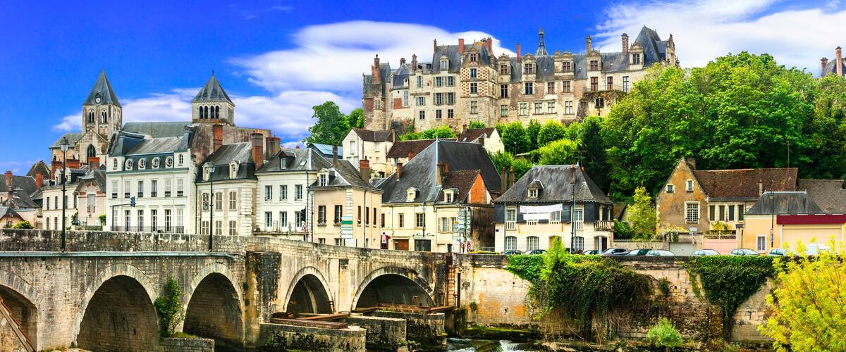 A historic town with a stone bridge, a prominent church tower, and a castle in the background.