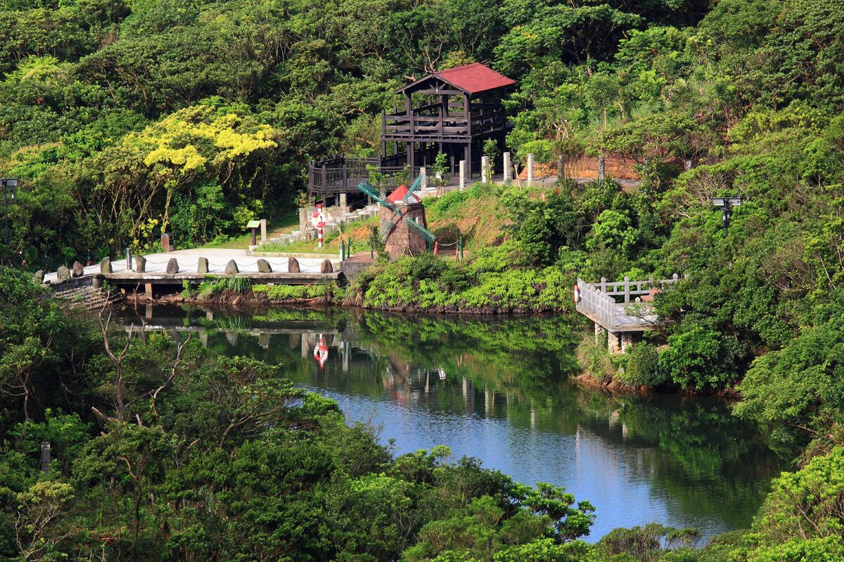 A serene lake surrounded by lush greenery, a traditional wooden structure, and a small bridge.