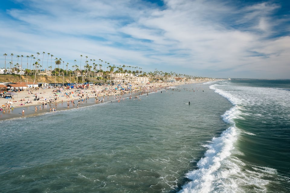 A beach with palm trees, people, and a lighthouse in the distance.