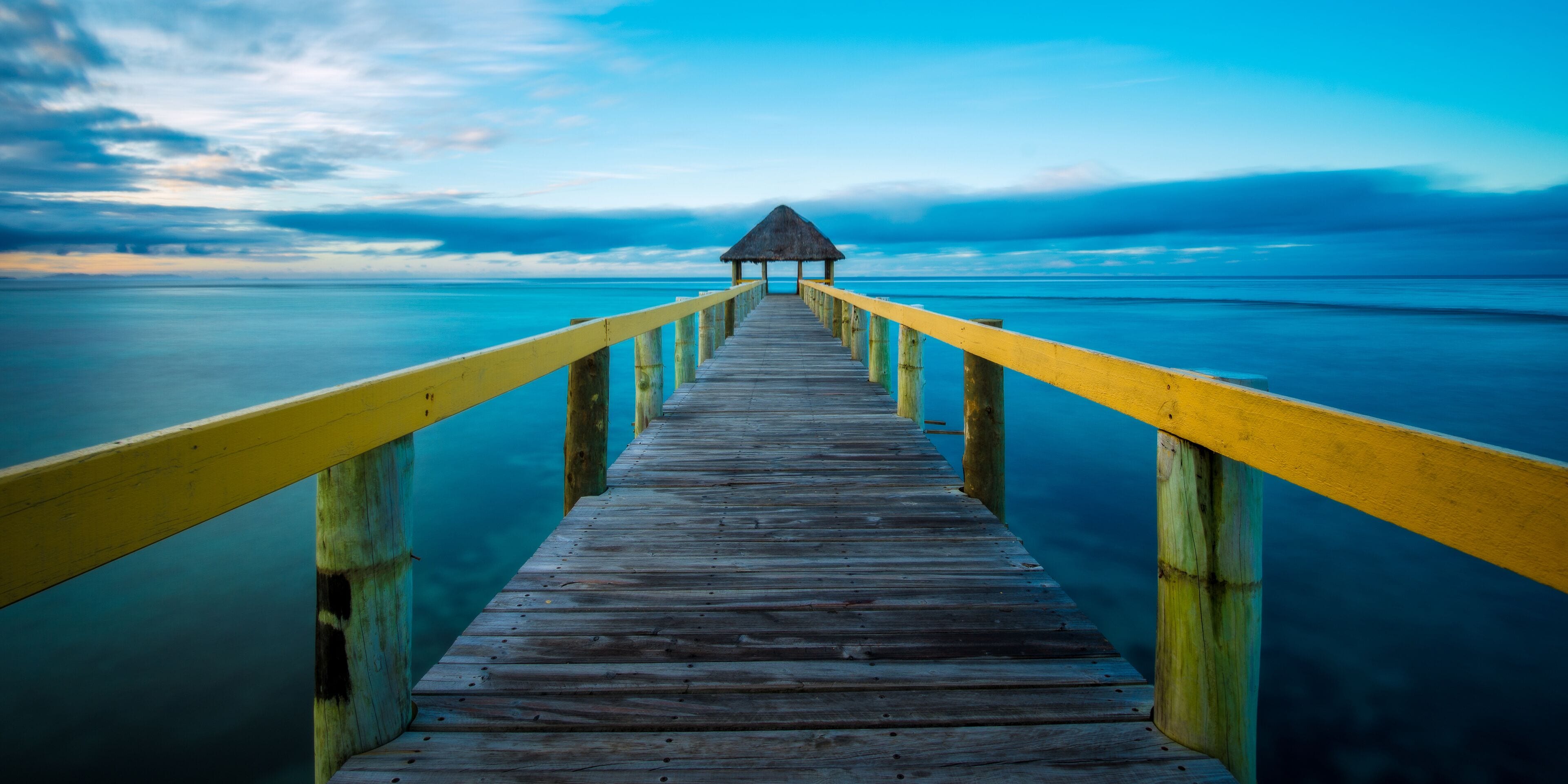 A wooden pier with yellow railings leading to a thatched-roof structure at the end.