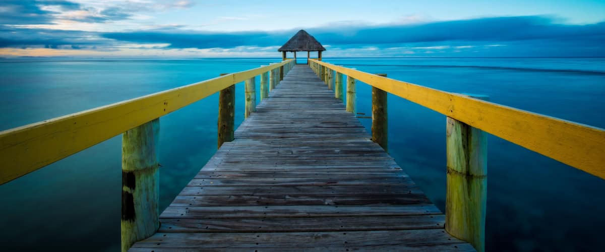 A wooden pier with yellow railings leading to a thatched-roof structure at the end.