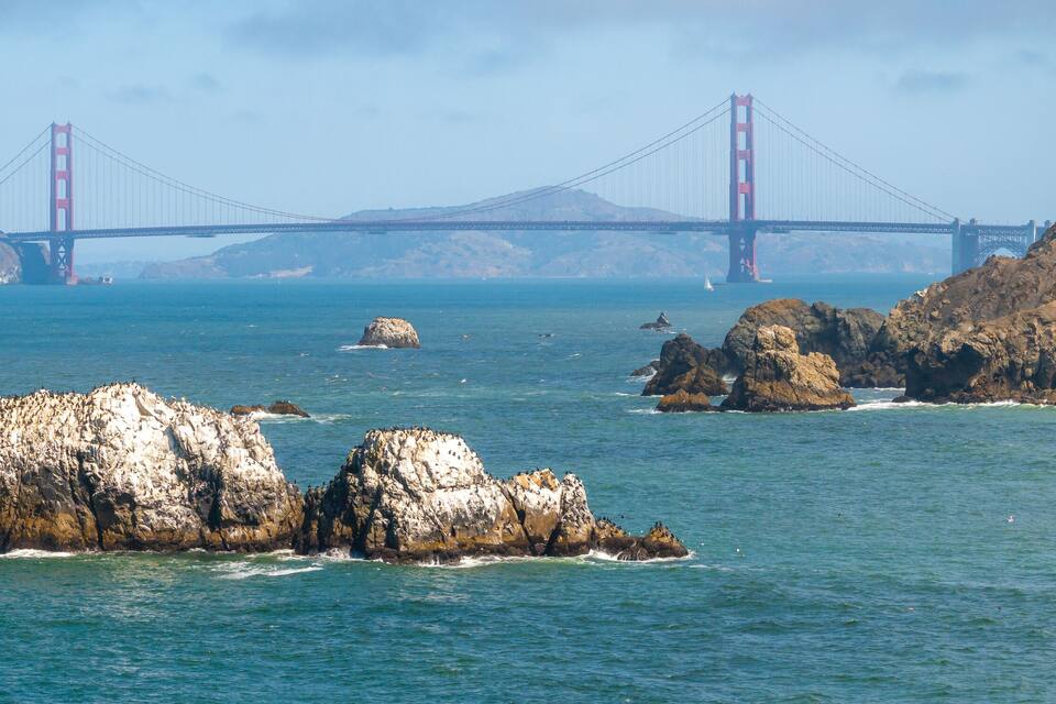 A coastal landscape with large rocks in the foreground and the Golden Gate Bridge in the background.