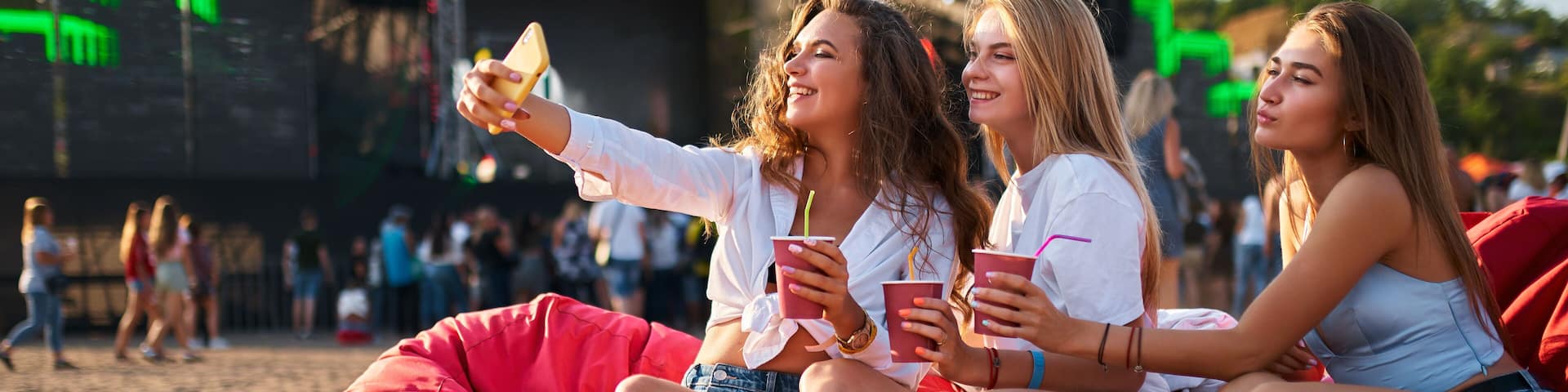 Girls relax on red bean bag chairs at beach music festival, enjoying drinks, toasting. Sunlit summer event with stage in background, youthful energy, leisure atmosphere. Women smiling, casual fashion.