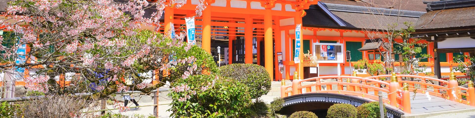 A traditional Japanese building with a bright orange structure, surrounded by cherry blossoms and a stone bridge.