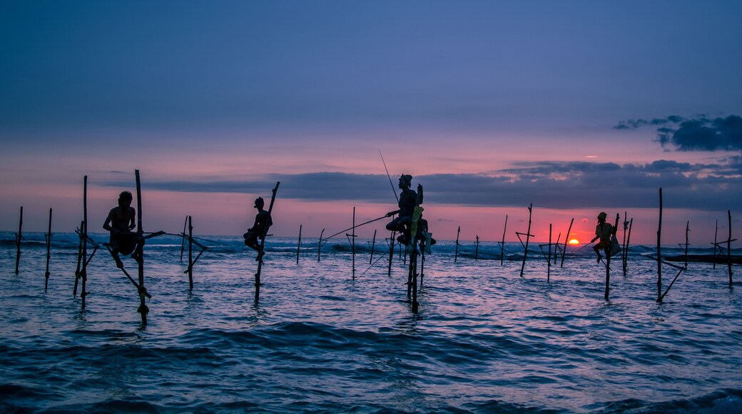 A sunset over the ocean with people fishing from bamboo poles.