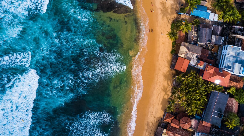 An aerial view of a beach with waves crashing onto the shore, surrounded by buildings and palm trees.