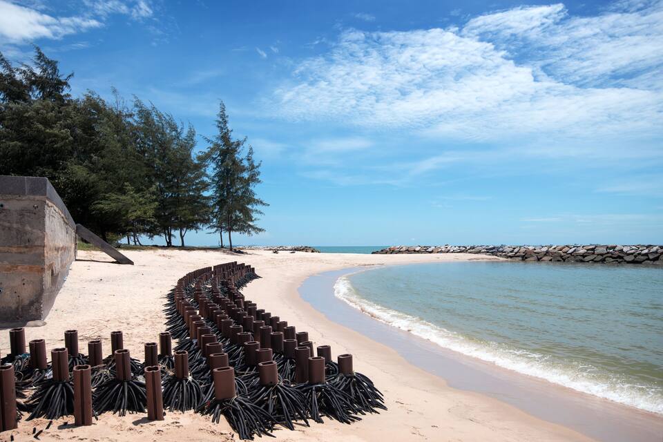 A sandy beach with a curved shoreline, a stone breakwater, and a row of cylindrical objects.