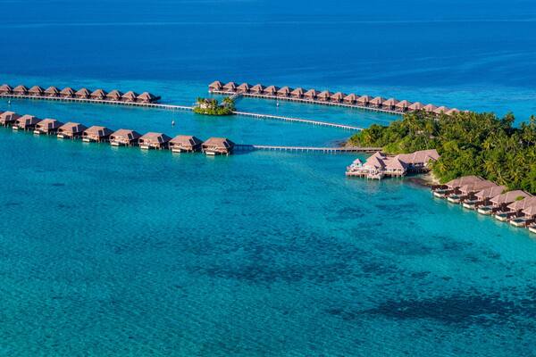 An aerial view of overwater bungalows connected by walkways, surrounded by turquoise waters and a tropical island.