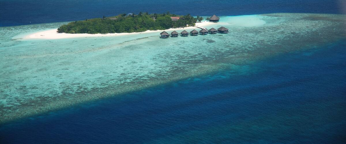 An island with overwater bungalows and a small building, surrounded by clear turquoise waters and a sandy beach.