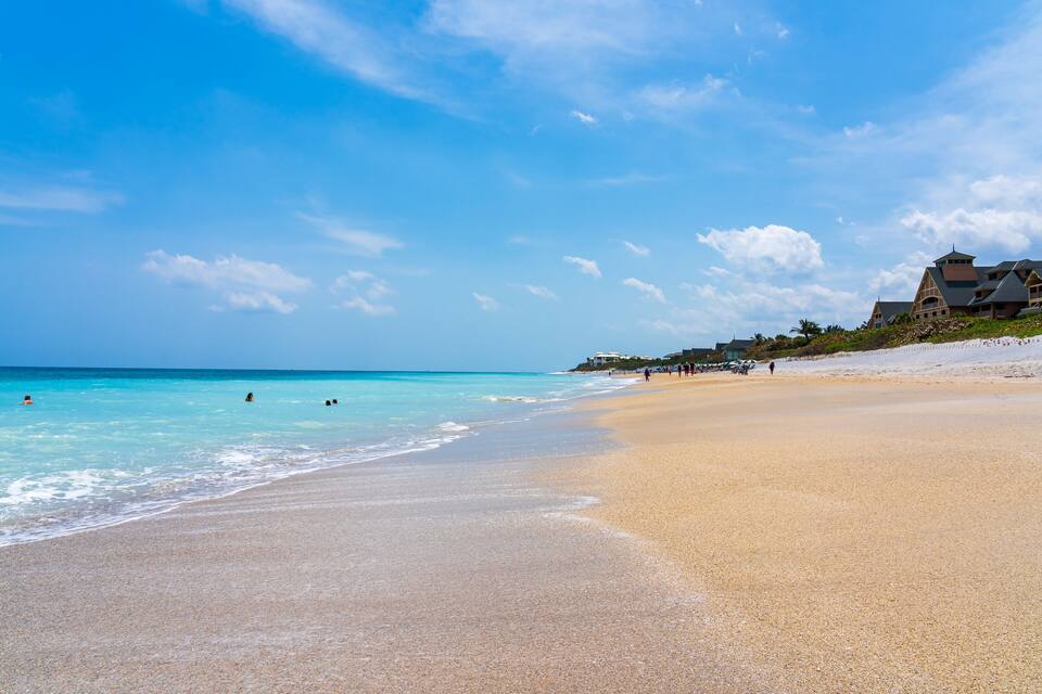 A beach with clear turquoise water, a sandy shore, and a resort building on the right.